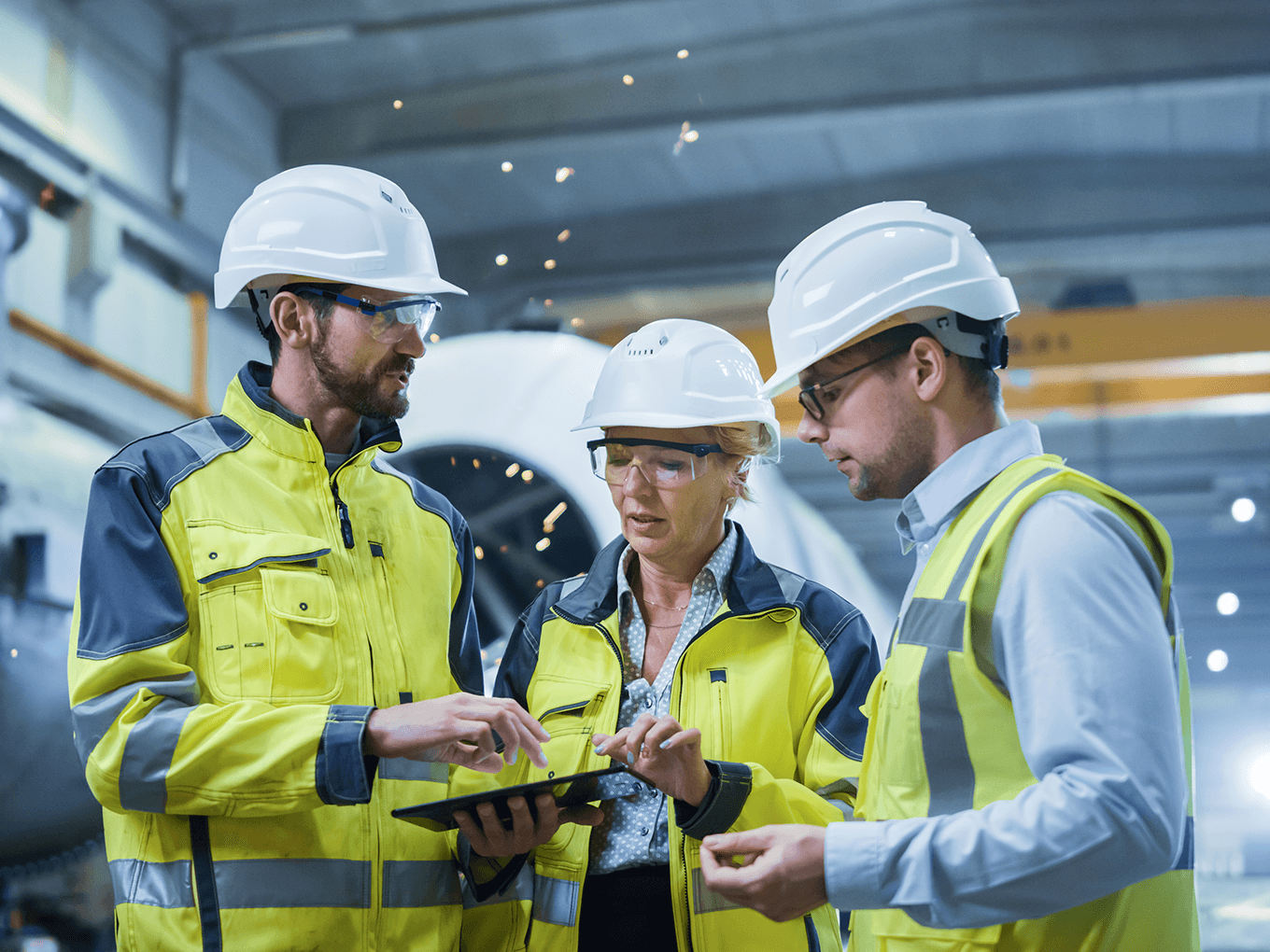 Drei Arbeitnehmer mit Helm und Schutzweste stehen in einer Produktionshalle vor einer großen Flugzeugturbine und besprechen sich.