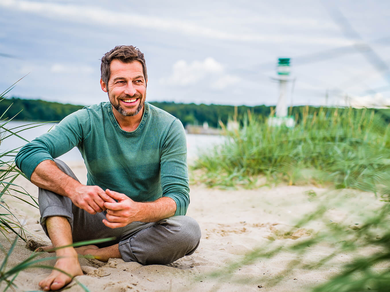 Ein Versicherungsmakler im Ruhestand sitzt entspannt in den Dünen, im Hintergrund ist ein Leuchtturm zu sehen.