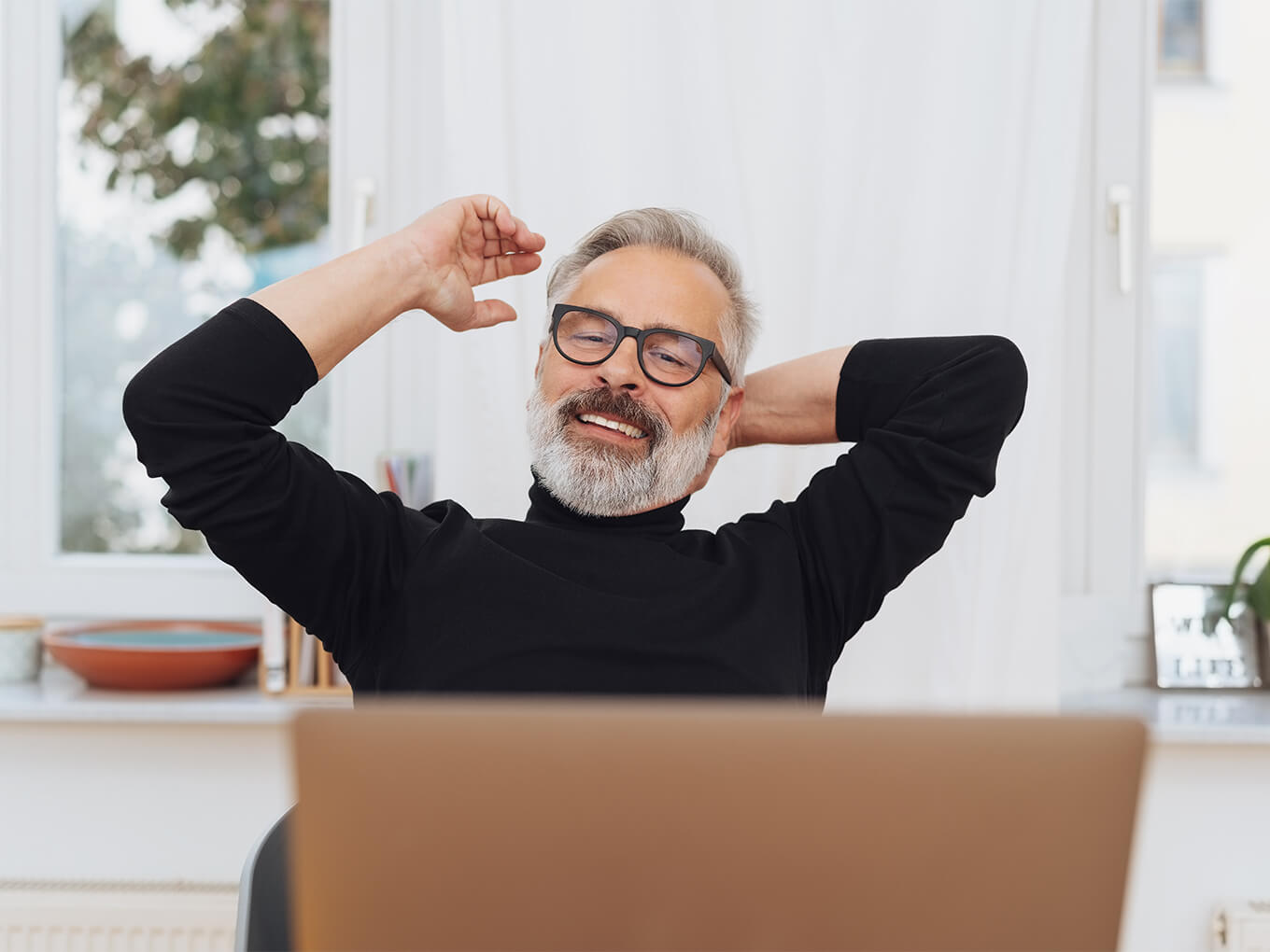 Ein älterer Versicherungsmakler mit grauem Bart sitzt entspannt vor seinem Notebook und streckt die Arme nach hinten.
