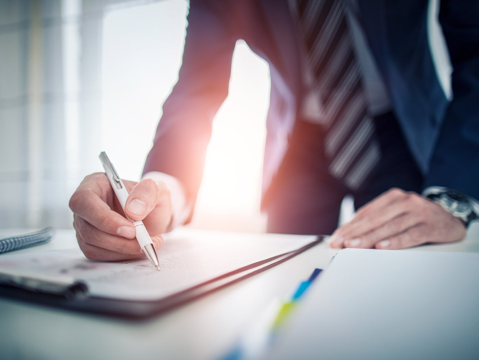 Eine Person im Business-Anzug schreibt mit einem Stift in ein Notizbuch auf einem Schreibtisch