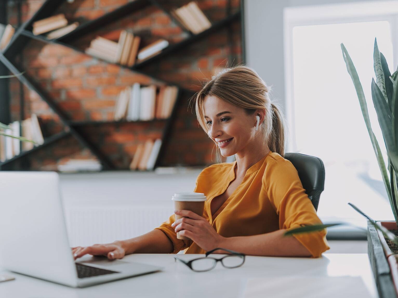 Versicherungsmaklerin sitzt entspannt mit einem Coffee-to-Go-Becher am Schreibtisch vor ihrem Notebook